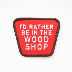 May include: Red fabric patch with brown stitching. The patch has white text that reads "I'd rather be in the wood shop."