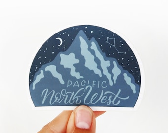 May include: A semi-circular sticker featuring a dark blue night sky with a crescent moon, a constellation, and light blue mountain peaks. The text Pacific North West is printed in white lettering across the lower portion of the mountain design.