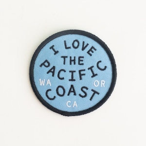 May include: A round, light blue patch with the text "I LOVE THE PACIFIC COAST" in black. The patch includes the abbreviations "WA" and "CA" and is bordered by a black stitched edge.