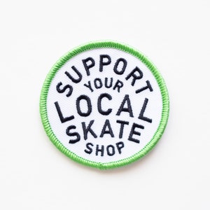 May include: White round patch with a green border. The patch has black embroidered text that reads "Support Your Local Skate Shop".