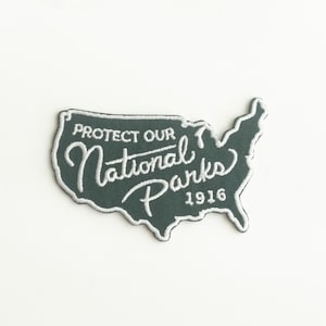 May include: A dark green embroidered patch in the shape of the United States. The patch features the text "PROTECT OUR National Parks 1916" in white script and block letters. The patch has a white border and is likely for a jacket or bag.