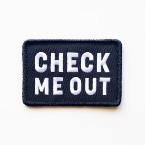 Peut inclure: Patch en tissu noir avec du texte brodé blanc qui dit "CHECK ME OUT".