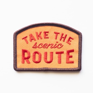 May include: A brown and orange embroidered patch with the text "Take the scenic route" in red.