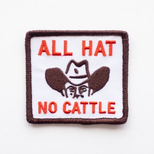 Peut inclure: Un écusson brodé marron et blanc avec le texte "ALL HAT NO CATTLE" en orange. Un dessin animé de cow-boy portant un chapeau marron est au centre de l'écusson.