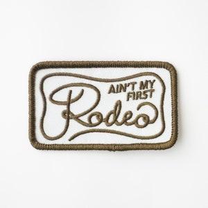 May include: A white embroidered patch with a brown border. The patch features the text "Ain't My First Rodeo" in a stylized font.