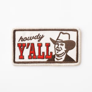 May include: A white embroidered patch with brown stitching. The patch features a cartoon cowboy wearing a brown hat and a red embroidered phrase that says "howdy y'all".