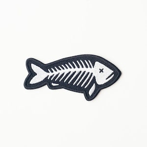 May include: Black embroidered patch of a fish skeleton with white stitching. The fish has an X over its eye.
