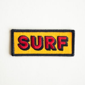 May include: A yellow embroidered patch with black stitching around the edges. The word "SURF" is embroidered in red with black outlines.