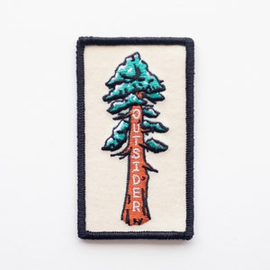 May include: An embroidered patch featuring a stylized green pine tree with a brown trunk. The word "OUTSIDER" is embroidered vertically on the trunk in white.