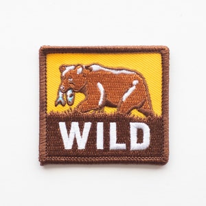 May include: Embroidered patch featuring a brown bear with a fish in its mouth, set against a yellow background. The word "WILD" is displayed in white letters on a brown base. The patch has a brown border.