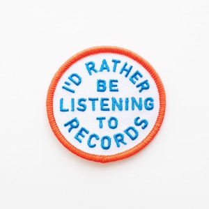 May include: White round patch with an orange border. The text "I'd rather be listening to records" is embroidered in blue.
