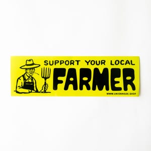 May include: A yellow bumper sticker with black text and a black and white illustration of a farmer. The text reads "SUPPORT YOUR LOCAL FARMER" and "www.unionmade.shop". The farmer is wearing overalls and a straw hat and holding a pitchfork.