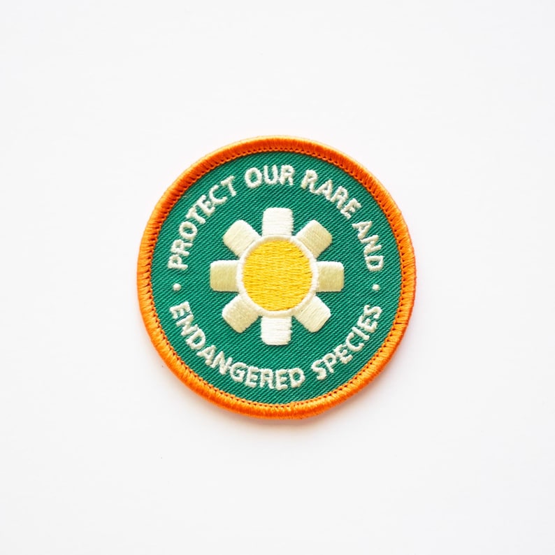 May include: Green and orange embroidered patch with a white flower and yellow centre. The text reads "Protect Our Rare and Endangered Species."