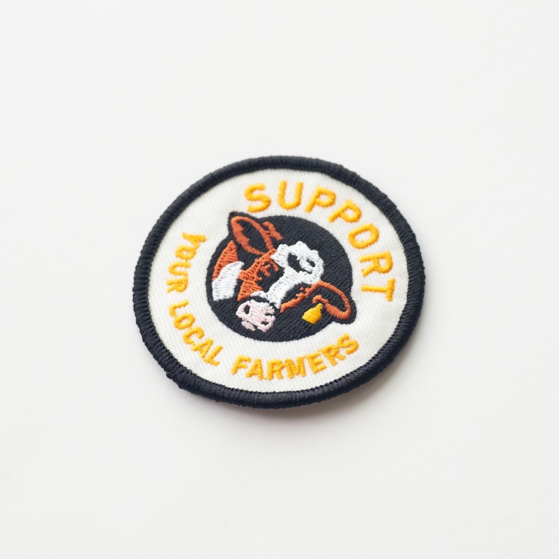 May include: A black and white embroidered patch with a cow's face in the center. The text around the cow says "Support Your Local Farmers" in yellow.