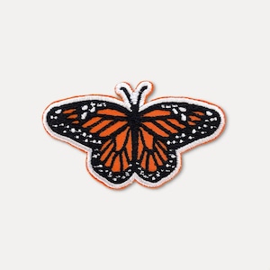 May include: Embroidered butterfly patch featuring orange and black wings with white dot details, outlined in black and white stitching. The patch has a white border and is designed to be ironed on or sewn onto fabric.
