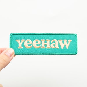 May include: A turquoise blue fabric patch with the word "yeehaw" embroidered in cream colored thread.