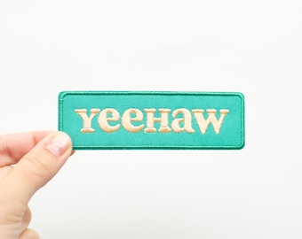May include: A turquoise blue fabric patch with the word "yeehaw" embroidered in cream coloured thread.