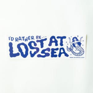 May include: A blue and white sticker with the text "I'd rather be... LOST AT SEA" in a playful font. A blue mermaid illustration is on the right. The sticker has a white background and the website address www.unionmade.shop.