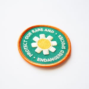 May include: A green embroidered patch with an orange border. The patch features a white daisy with a yellow centre and the text "PROTECT OUR RARE AND ENDANGERED SPECIES".