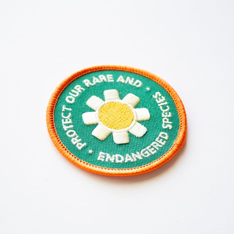 May include: A green embroidered patch with an orange border. The patch features a white daisy with a yellow centre and the text "PROTECT OUR RARE AND ENDANGERED SPECIES".
