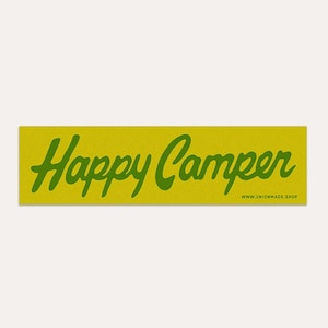 Op de afbeelding: Een rechthoekige sticker met de woorden "Happy Camper" in groene cursieve letters op een gele achtergrond. Het websiteadres www.unionmade.shop staat onder de tekst afgedrukt.