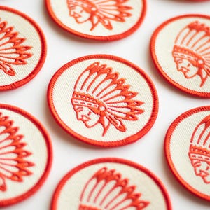 K&ouml;nnte beinhalten: Sechs gestickte B&uuml;gelbilder mit einem wei&szlig;en indianischen Kopfschmuck-Design auf cremefarbenem Hintergrund mit rotem Rand.