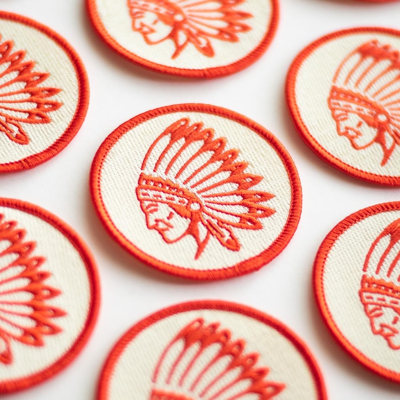 K&ouml;nnte beinhalten: Sechs gestickte B&uuml;gelbilder mit einem wei&szlig;en indianischen Kopfschmuck-Design auf cremefarbenem Hintergrund mit rotem Rand.