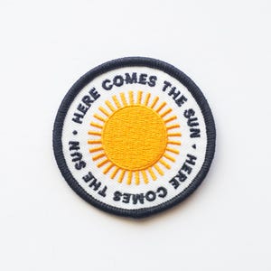 May include: A white embroidered patch with a black border. The patch features a yellow sun with rays and the text "Here Comes The Sun. Here Comes The Sun."