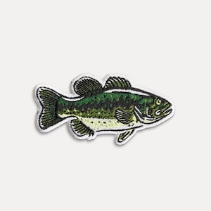 May include: Embroidered patch featuring a detailed illustration of a green and black fish with white accents. The fish has a textured pattern and is outlined with white stitching. This patch is suitable for adding to clothing or accessories.