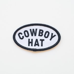 May include: An oval patch featuring the text "COWBOY HAT" in black, embroidered on a white background. The patch has a black border. It is designed to be sewn onto clothing or accessories, such as a jacket or bag.