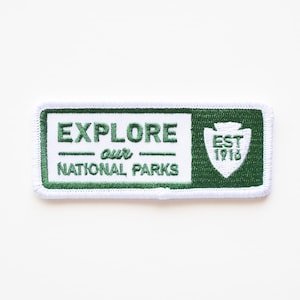 May include: White embroidered patch with green text and border. The text reads "EXPLORE our NATIONAL PARKS" with a green arrowhead design to the right that says "EST 1916".