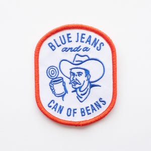 May include: Blue embroidered patch with a cowboy drinking from a can of beans. The text reads "Blue Jeans and a Can of Beans".