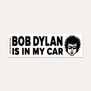 May include: A rectangular white bumper sticker with black text that reads "BOB DYLAN IS IN MY CAR." A black and white illustration of Bob Dylan with sunglasses and curly hair is on the right side. The website address www.unionmade.shop is on the left side.