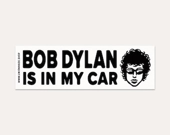May include: A rectangular white bumper sticker with black text that reads "BOB DYLAN IS IN MY CAR." A black and white illustration of Bob Dylan with sunglasses and curly hair is on the right side. The website address www.unionmade.shop is on the left side.