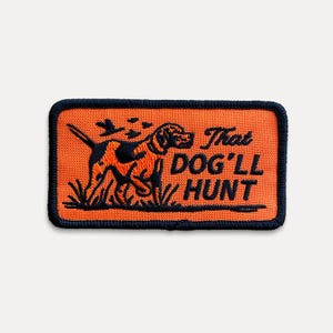 May include: Embroidered patch with a rectangular shape. The patch features an orange background with a black border. A dog illustration is in the center, with the text "That DOG'LL HUNT" below the dog. The dog is black and brown.