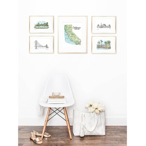 May include: A collection of framed art prints featuring California landmarks and a map of the state. The prints are in gold frames and hang above a white chair, a tote bag with flowers, and gold sandals.