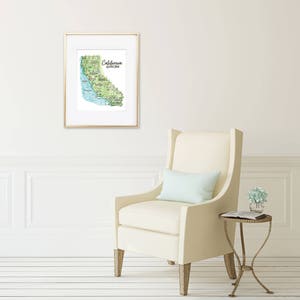 May include: A framed watercolor map of California, with green, blue, and beige tones. The map is in a gold frame and hangs on a white wall. The text "California Golden State" is visible. A cream-colored armchair and side table are in the foreground.