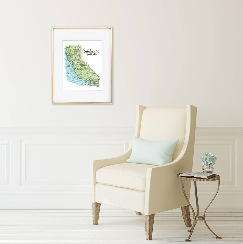 May include: A framed watercolor map of California, with green, blue, and beige tones. The map is in a gold frame and hangs on a white wall. The text "California Golden State" is visible. A cream-colored armchair and side table are in the foreground.
