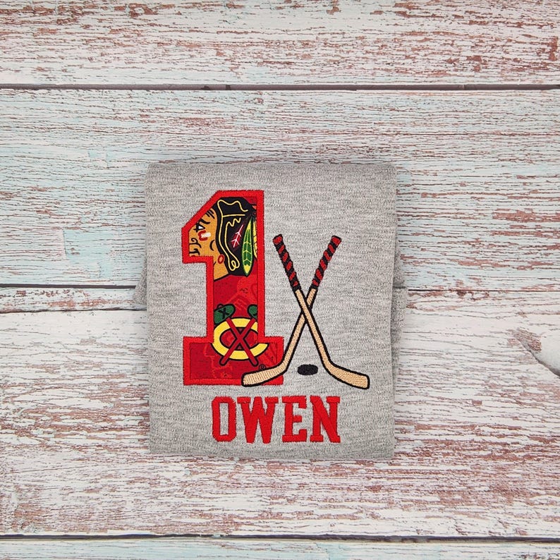 May include: A gray t-shirt with a red embroidered number 1, a hockey stick design, and the name "OWEN" in red. The number 1 features a Native American head logo and a hockey puck.