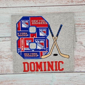 May include: A gray t-shirt featuring a large embroidered number 8 in blue, red, and white, with "NEW YORK RANGERS" text. Crossed hockey sticks and the name "DOMINIC" are also embroidered in red.