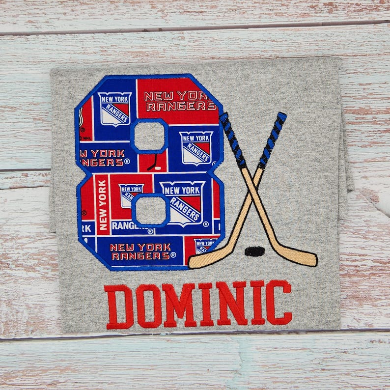 May include: A gray t-shirt featuring a large embroidered number 8 in blue, red, and white, with "NEW YORK RANGERS" text. Crossed hockey sticks and the name "DOMINIC" are also embroidered in red.