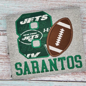 May include: Gray t-shirt with a green and white New York Jets logo, the number 8, and a brown football. The name "SARANTOS" is embroidered in green below the logo.