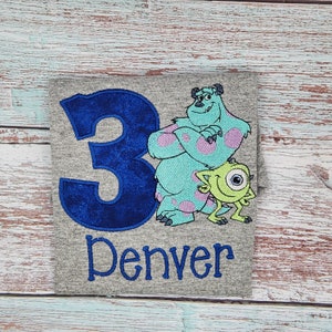 May include: A gray t-shirt with a blue number 3, a blue and green monster character, and a green one-eyed monster character. The text "Denver" is embroidered below the characters.