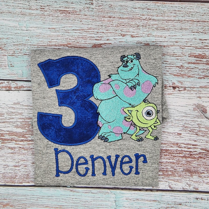 May include: A gray t-shirt with a blue number 3, a blue and green monster character, and a green one-eyed monster character. The text "Denver" is embroidered below the characters.
