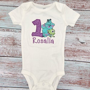 May include: White onesie with a purple glitter number one, a blue and green monster character, and the name "Rosalia" embroidered in purple.