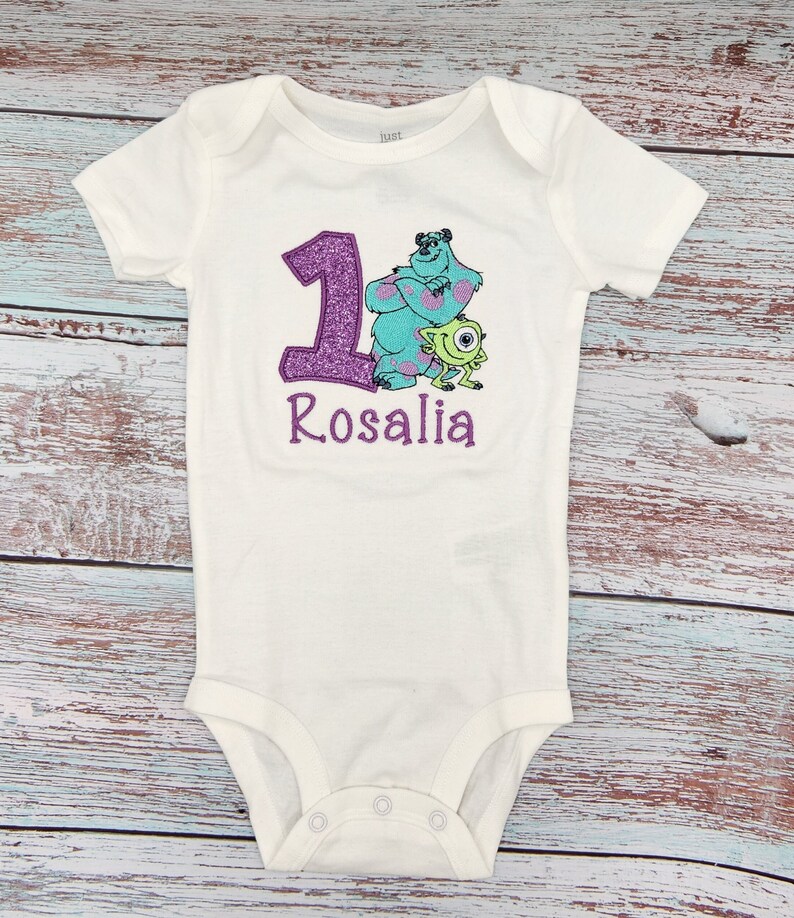 May include: White onesie with a purple glitter number one, a blue and green monster character, and the name "Rosalia" embroidered in purple.