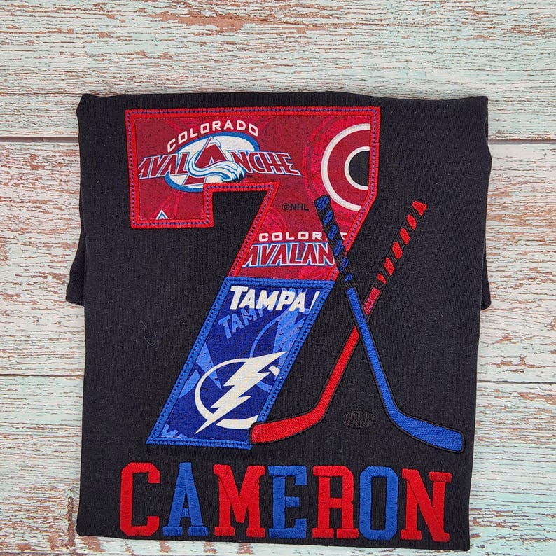 May include: Black t-shirt featuring hockey-themed graphics. The design includes the Colorado Avalanche and Tampa Bay Lightning logos, along with crossed hockey sticks. The name "CAMERON" is embroidered in red and blue.