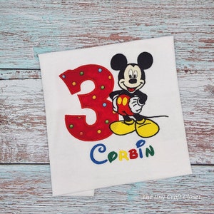 May include: White t-shirt with red, blue, yellow, and green embroidered Mickey Mouse, the number 3, and the name Corbin.
