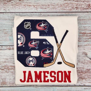 May include: White t-shirt with a large embroidered number 6 in navy blue, featuring Columbus Blue Jackets logos and the NHL logo. Crossed hockey sticks and the name "JAMESON" in red complete the design. A puck is also visible.