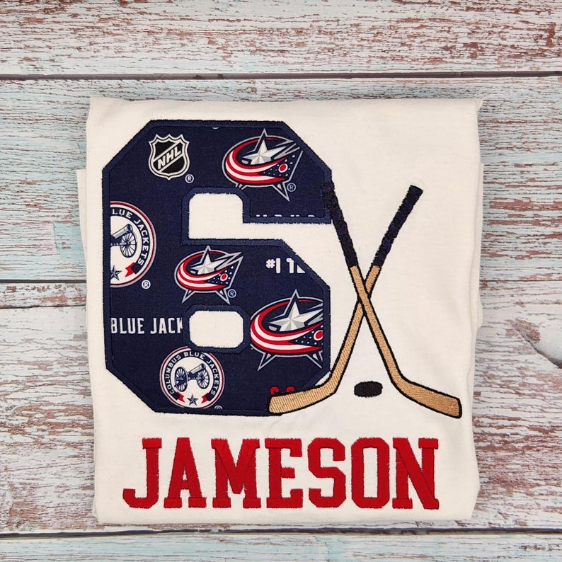 May include: White t-shirt with a large embroidered number 6 in navy blue, featuring Columbus Blue Jackets logos and the NHL logo. Crossed hockey sticks and the name "JAMESON" in red complete the design. A puck is also visible.